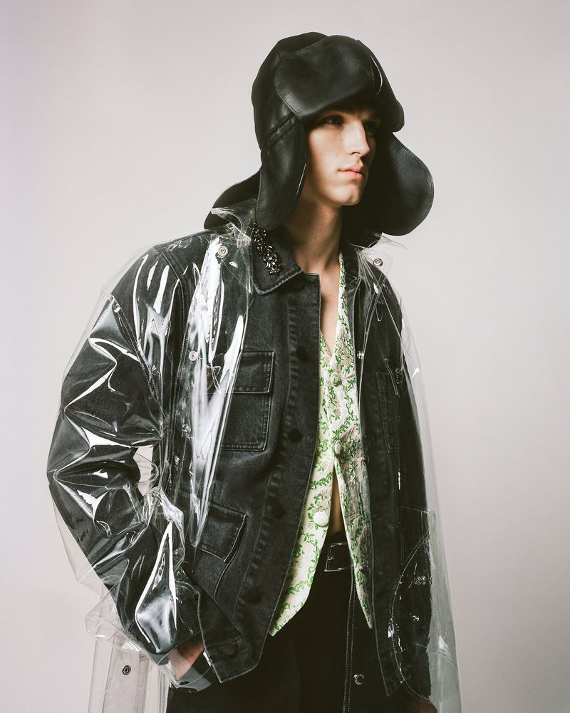 Male model poses in a transparent vinyl coat, black denim jacket, and black pants, complemented by a distinctive black hat.