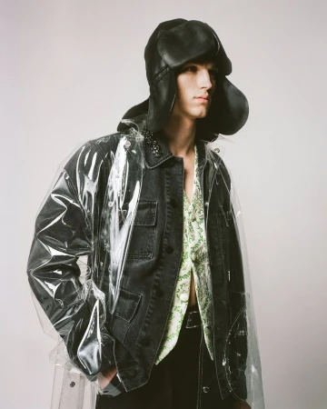 Male model poses in a transparent vinyl coat, black denim jacket, and black pants, complemented by a distinctive black hat.