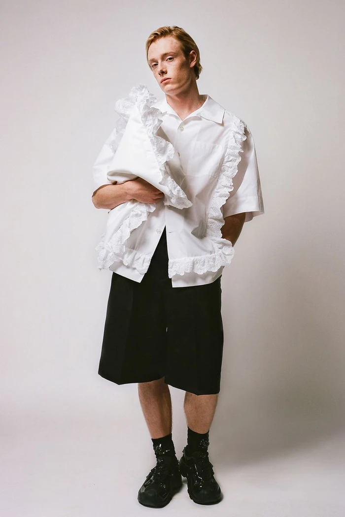 Model in white ruffled shirt and black shorts against a plain light studio background