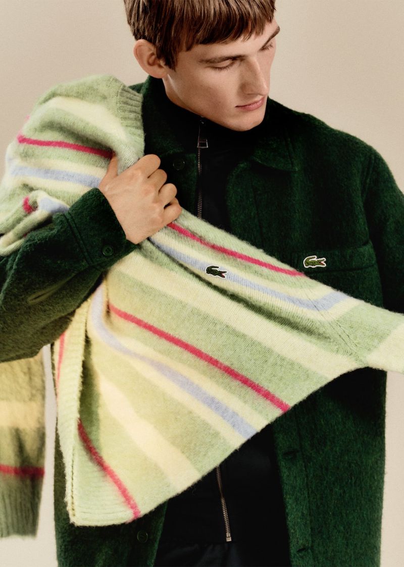 Man poses in a green Lacoste jacket and sweater in a studio setting, showcasing fashion apparel with the brand's iconic logo.