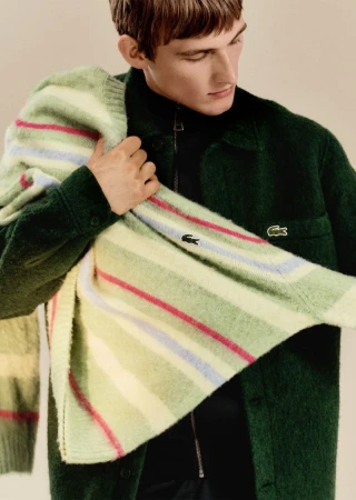 Man poses in a green Lacoste jacket and sweater in a studio setting, showcasing fashion apparel with the brand's iconic logo.