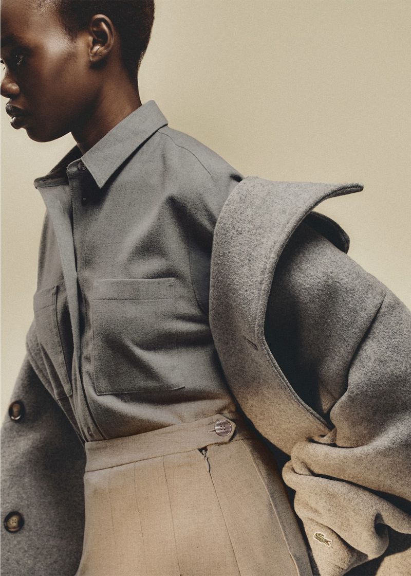 Fashion shot of a Black model wearing a grey button-down shirt with a jacket draped over the shoulder, and a khaki skirt.