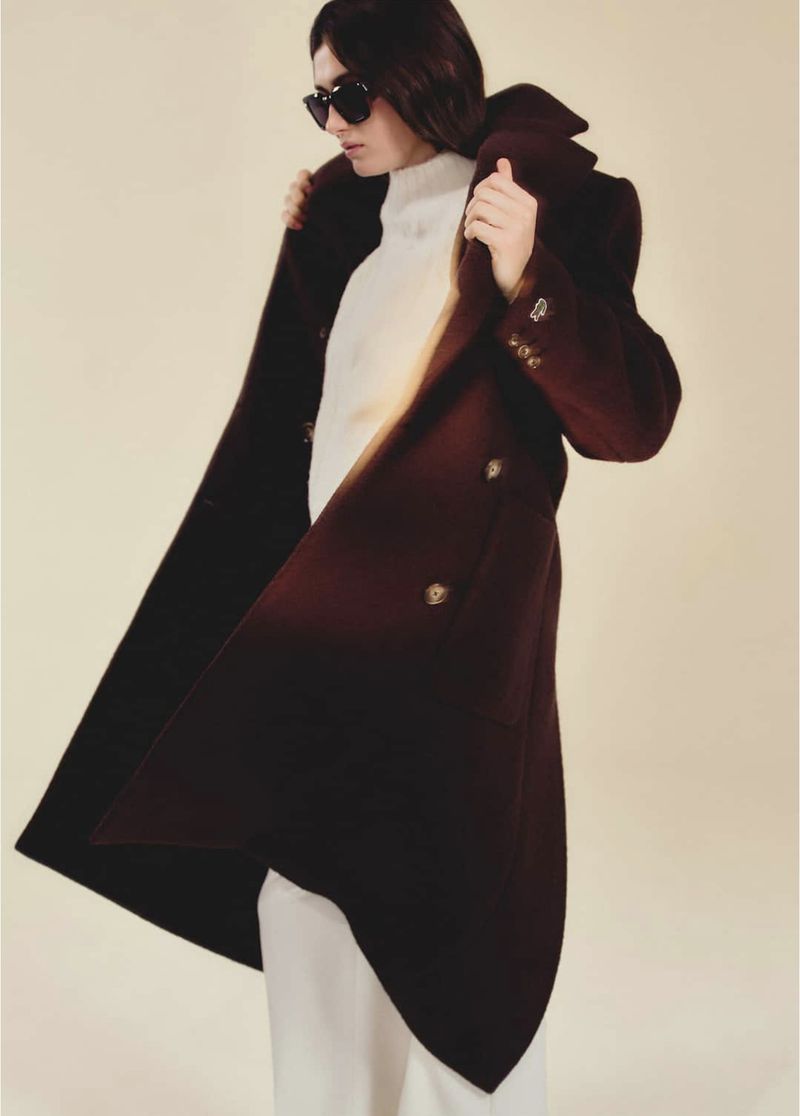 A woman in sunglasses and a long brown coat over a white sweater and pants poses in a studio with a neutral background.