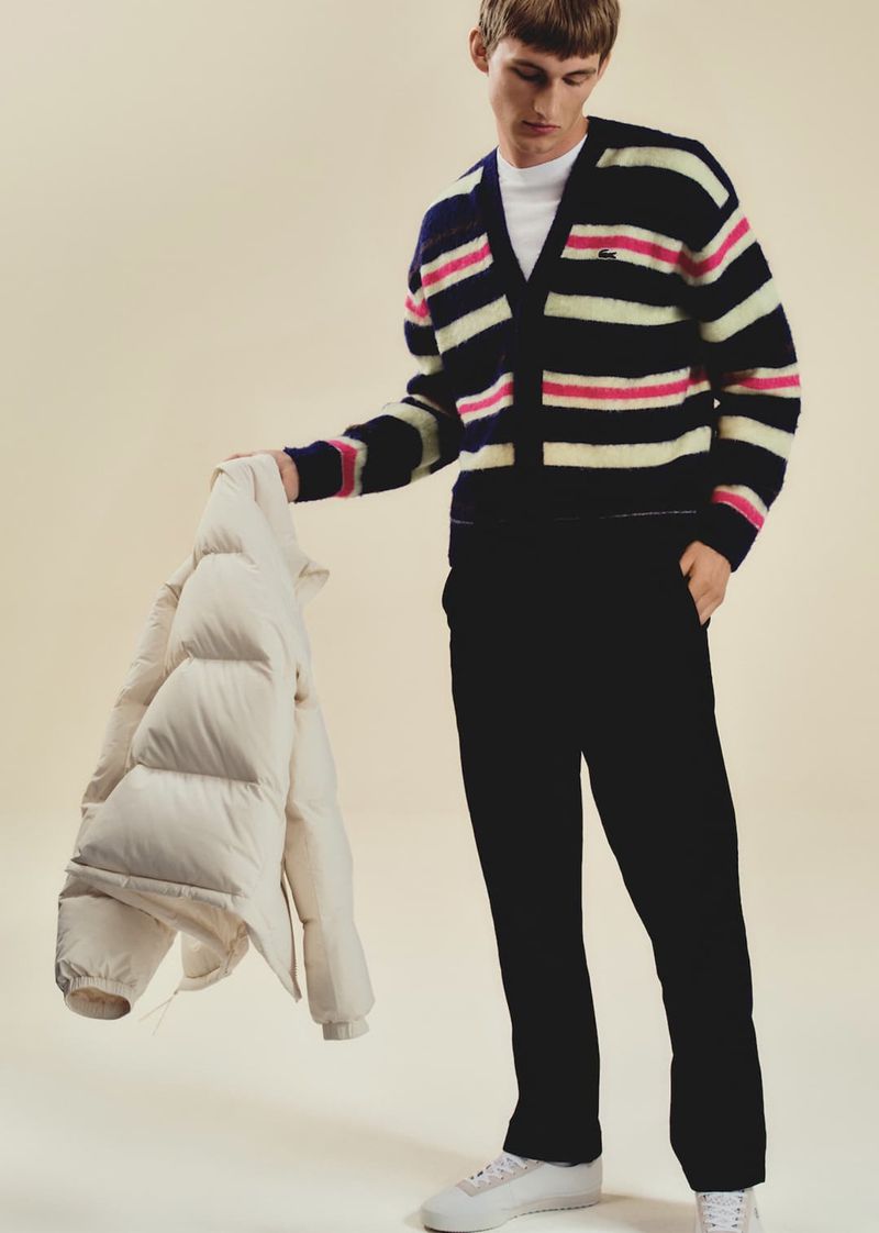 Male fashion model poses in a striped cardigan, holding a puffer jacket, standing in a studio setting with a neutral background.