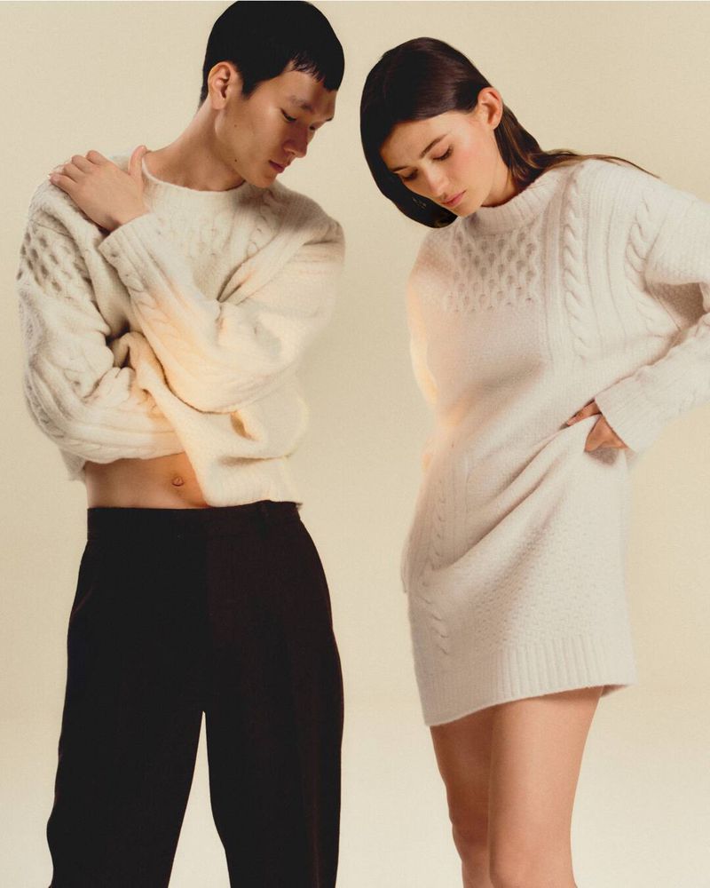 Two fashion models showcase knitted sweaters indoors. An adult Asian male poses alongside a Caucasian woman in a cream-color backdrop.