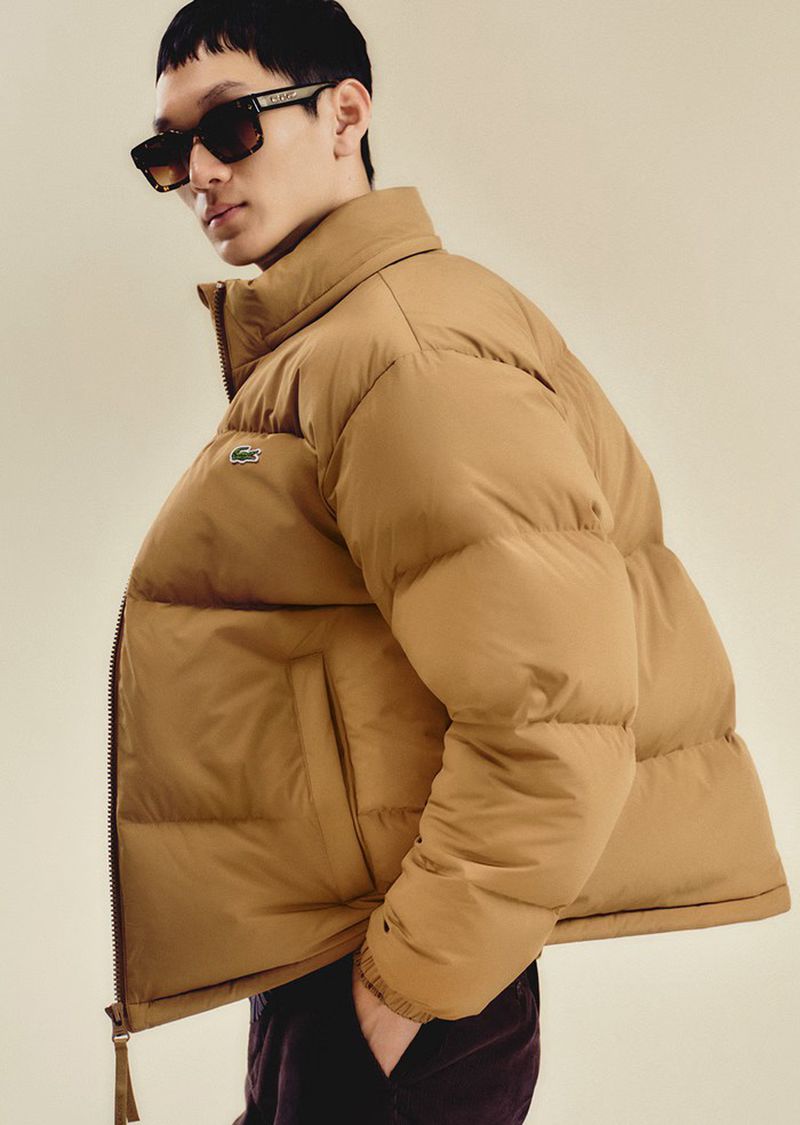 A slender man wearing a tan puffer jacket and sunglasses stands indoors against a muted backdrop, showcasing a modern yet casual look.