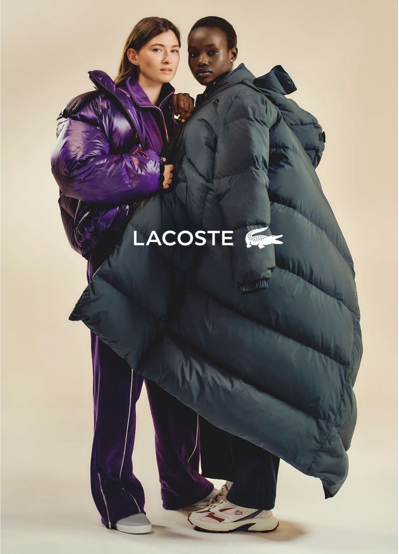 Two diverse women model oversized Lacoste puffer coats in a fashion shot. One wears purple while the other sports a navy blue coat.