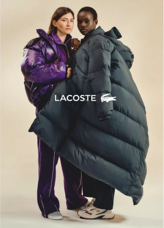 Two diverse women model oversized Lacoste puffer coats in a fashion shot. One wears purple while the other sports a navy blue coat.
