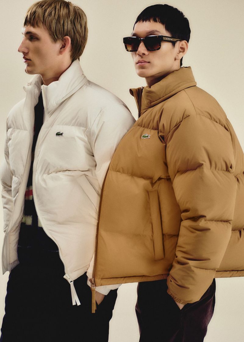 Two men pose in tan and cream Lacoste puffer jackets against a neutral backdrop, one wearing sunglasses, showcasing a fashion-forward style.