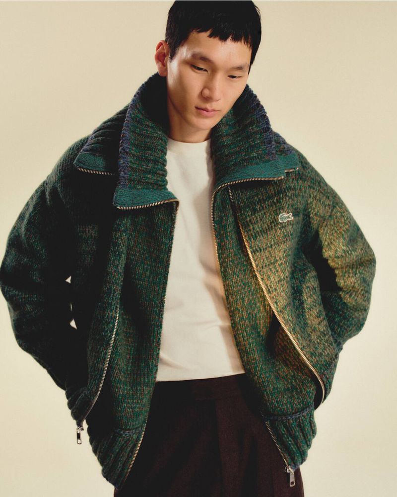 Asian male model poses in a green knitted Lacoste jacket, a white t-shirt, and brown pants in a neutral studio setting.