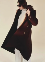 Model in dark sunglasses wearing a dark brown winter coat and white turtleneck sweater against a warm neutral background