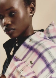 Close-up portrait of a model wearing a pink and white plaid jacket with a small embroidered crocodile logo for Lacoste.