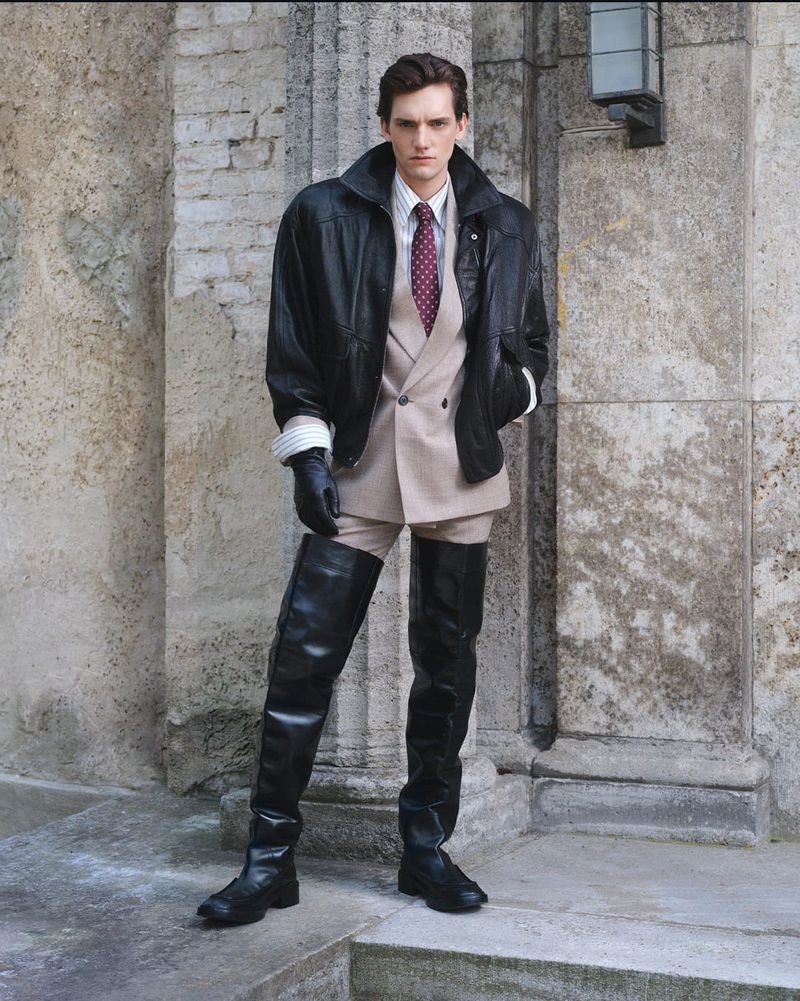 Fashionable male model posing in high-fashion black leather thigh-high boots, jacket, and beige suit, standing against a weathered stone wall.