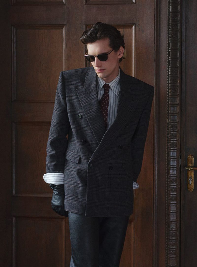 Man in a grey plaid suit with sunglasses and black gloves, posing against a dark wooden door for a fashion shoot in a studio.