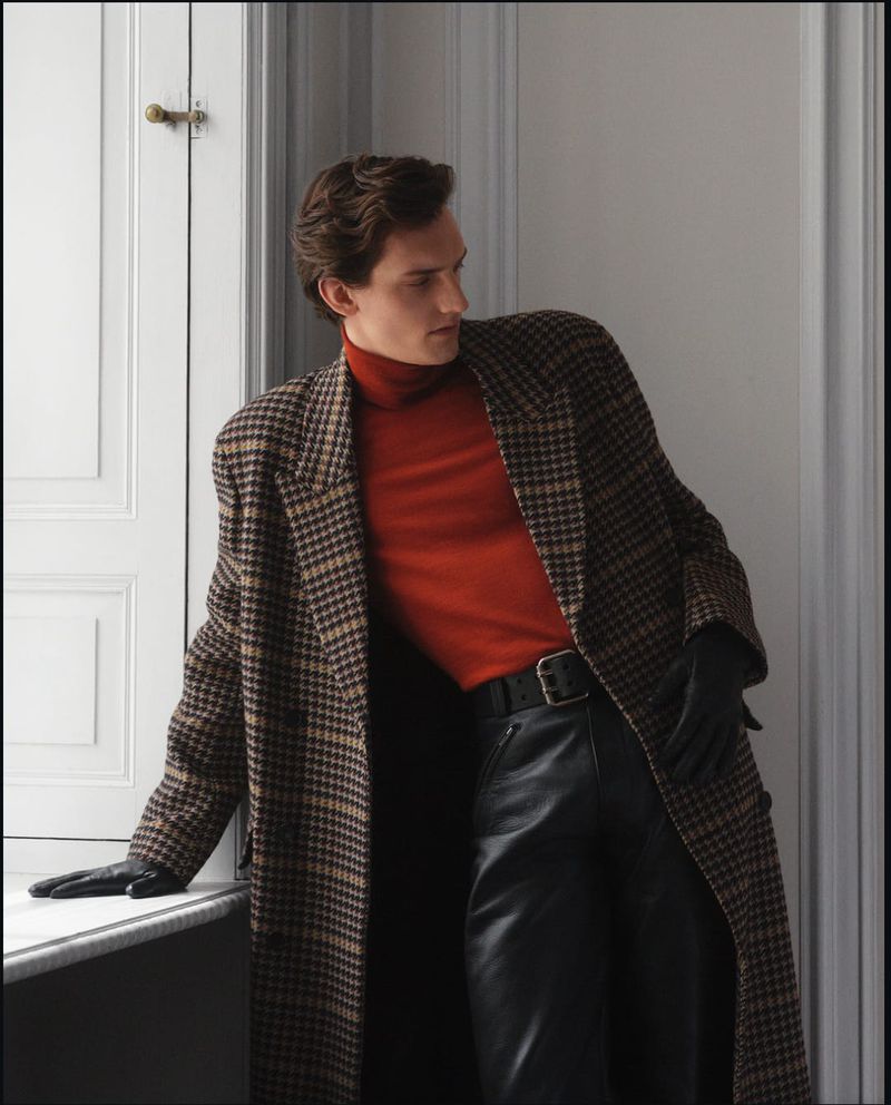 A stylish adult male leans against a window wearing a brown plaid overcoat, red turtleneck sweater, black leather pants, and black leather gloves.