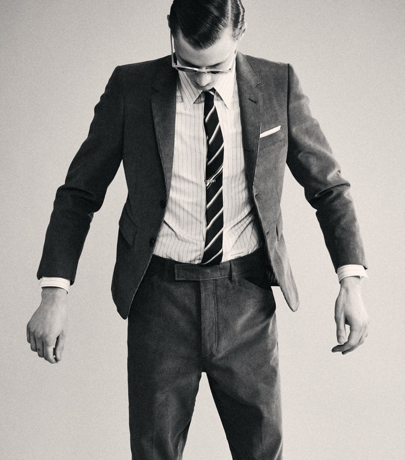 Monochrome studio shot of a man in a grey suit, vertically striped shirt, and diagonally striped tie, looking down with his arms hanging.