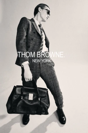 Man in tweed suit holding black leather satchel, wears sunglasses, posing in a fashion shot for Thom Browne, New York.