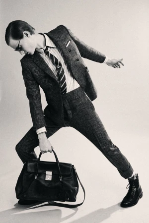 Model in plaid suit holding a black leather handbag against a light background for Thom Browne Fall 25