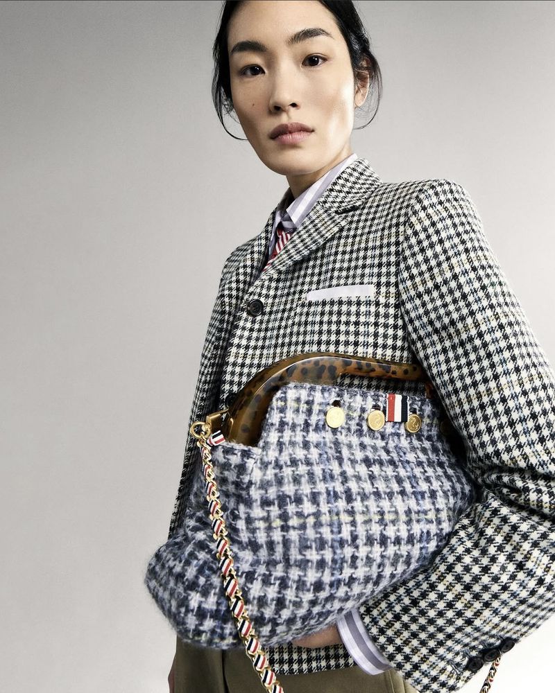 Asian fashion model poses indoors holding a tweed purse with a black-and-white houndstooth blazer and a neutral background in a portrait shot.