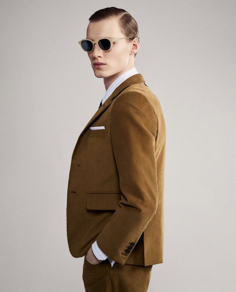 Portrait of an adult male model in a tan corduroy suit and sunglasses, posing in a studio with a plain, neutral background.