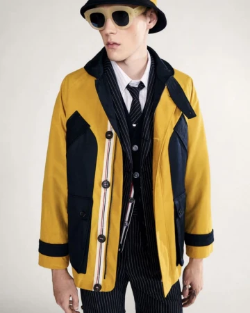 Fashionable man in yellow and black jacket, pinstripe suit, bucket hat, and sunglasses stands against a neutral backdrop in a studio.