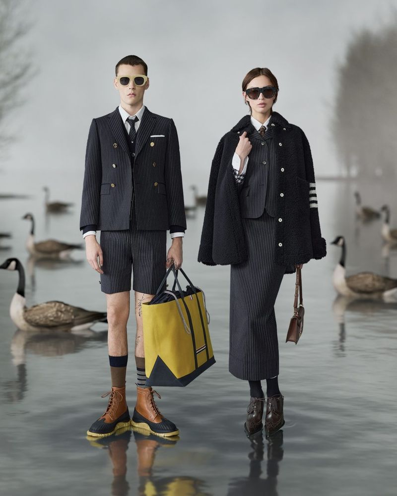 Two fashion models pose in pinstripe suits and sunglasses on water, holding bags, with geese in the background on a misty day.