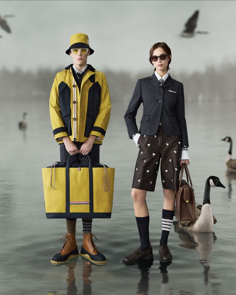Two fashion models pose outdoors in Thom Browne attire: a yellow rain jacket and tote bag, and a pinstripe blazer with brown shorts and tote.