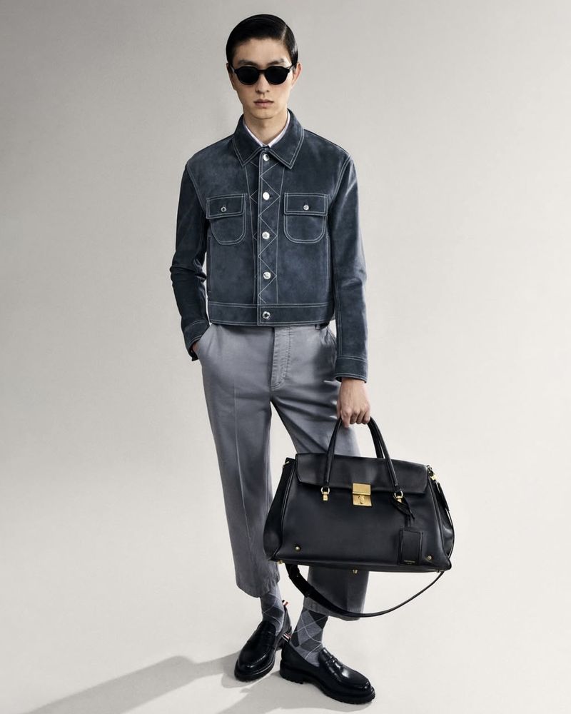 Male model in a grey suede jacket and trousers holding a black leather handbag against a plain background