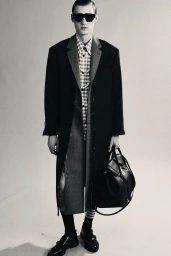 Model in dark sunglasses and black coat wearing a patterned shirt, holding a large bag against a light background