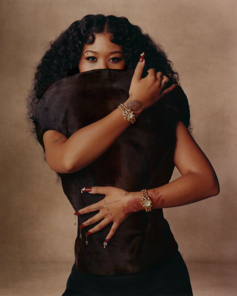Portrait of a woman with dark curly hair, adorned with ornate jewelry, partially obscured by a dark fur-like garment against a neutral backdrop.