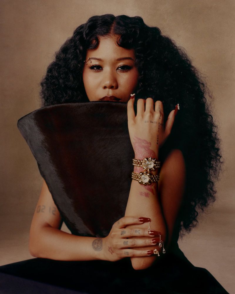 Portrait of a stylish Asian woman with curly hair, jewelry, dark dress, and a triangular object, set against a neutral background.