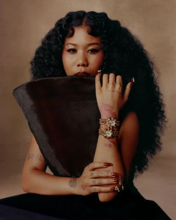 Portrait of a stylish Asian woman with curly hair, jewelry, dark dress, and a triangular object, set against a neutral background.