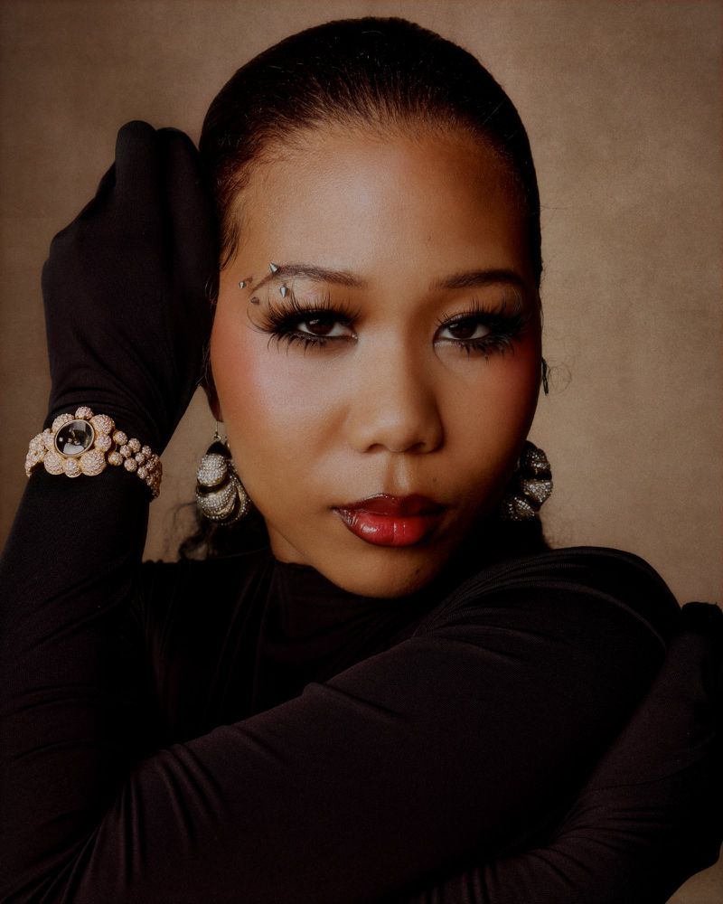 Portrait of a woman wearing black gloves, pearl bracelet, and textured earrings against a neutral textured background.