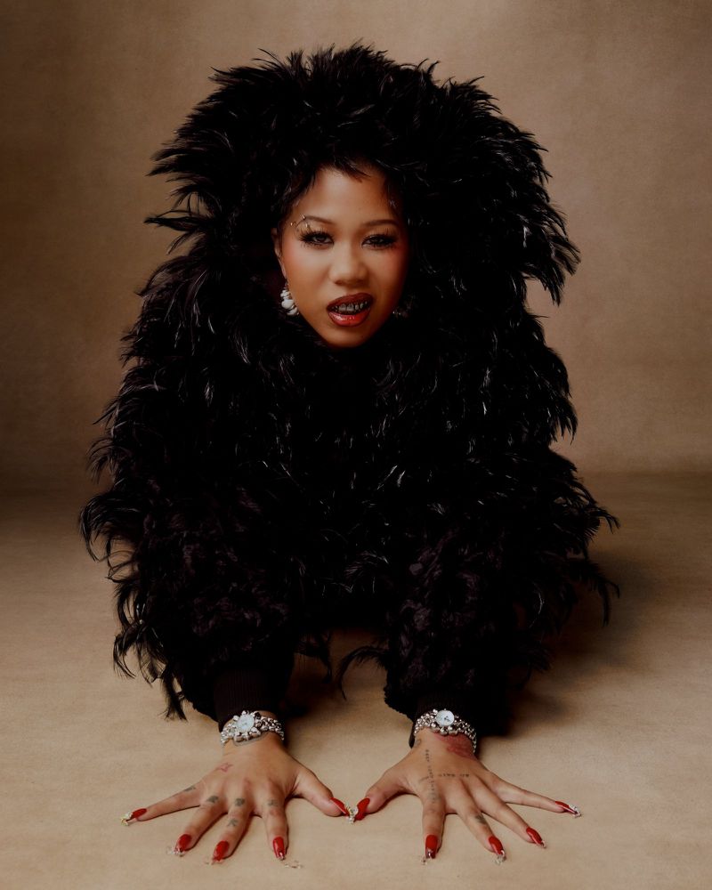 Portrait of a woman kneeling in a feathered boa with long red nails and jewelery on a beige background in a studio photoshoot.
