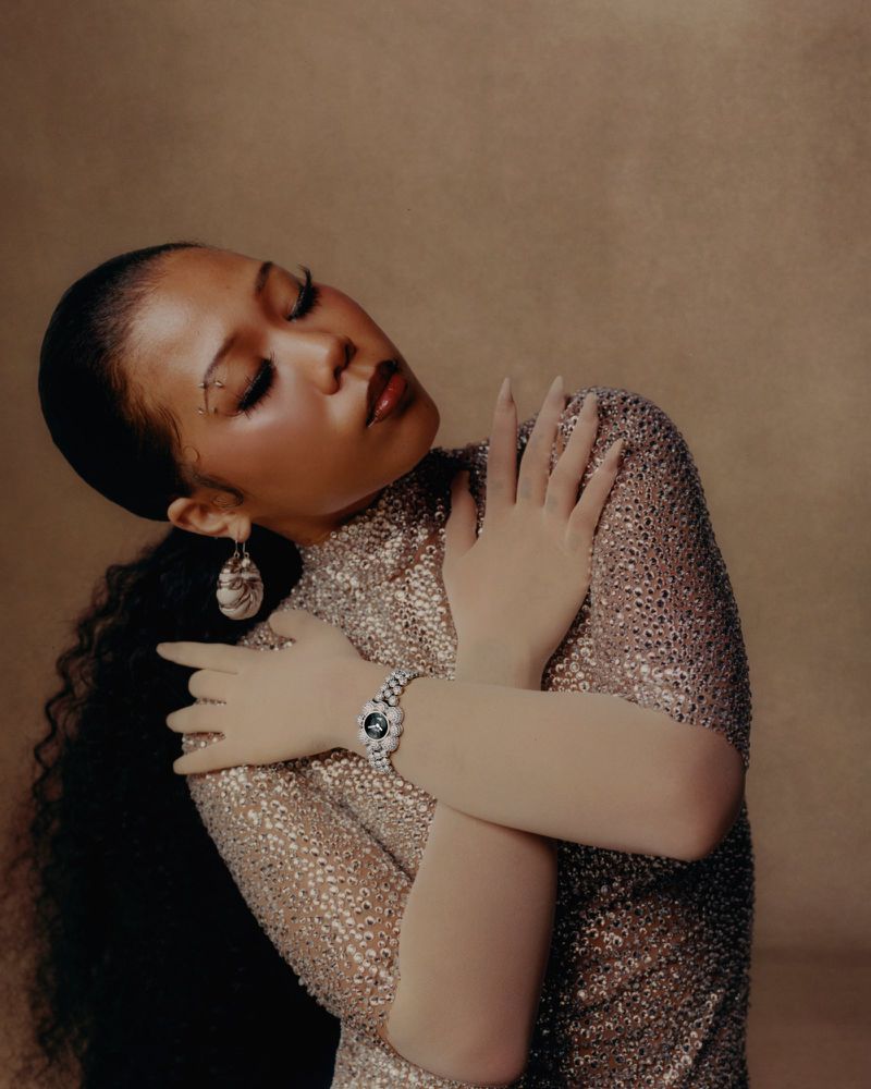 Portrait of a woman with dark hair and glitter makeup, wearing a sparkling dress and a watch with flower embellishments in a studio.