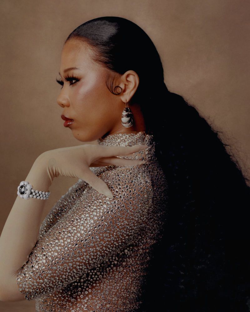 Profile portrait of a woman wearing a sparkling embellished high-neck top and pearl jewelry against a warm textured background.
