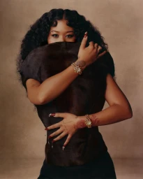 Close-up portrait of a Black woman with dark curly hair covering the lower half of her face, wearing gold watches and dark top