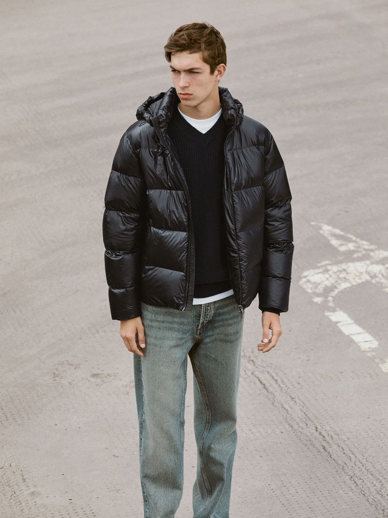A young man in a black puffer jacket, dark sweater, and light jeans stands on pavement. Portrait-style fashion shot with neutral background.