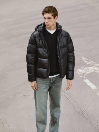 A young man in a black puffer jacket, dark sweater, and light jeans stands on pavement. Portrait-style fashion shot with neutral background.