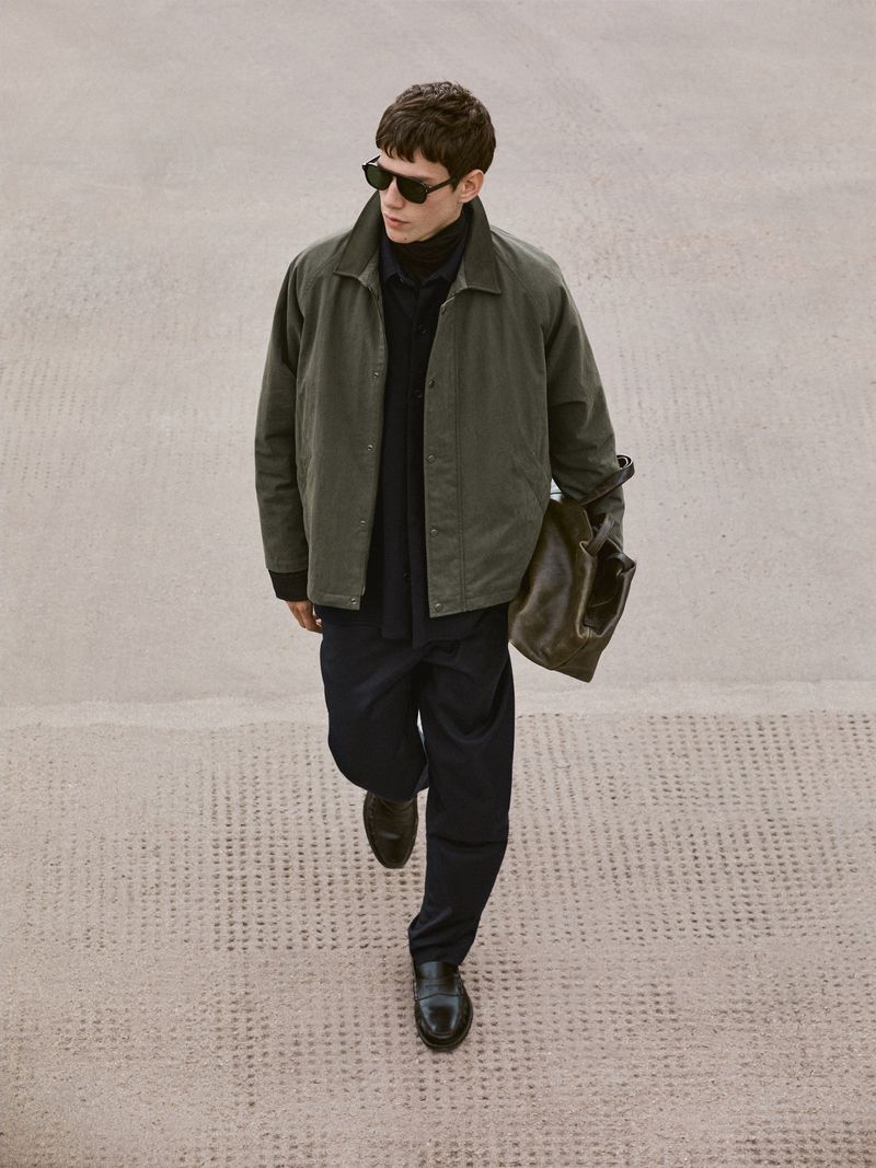 Man in olive jacket, navy pants, and sunglasses walks on concrete holding a tote bag, showcasing a blend of casual and sophisticated style.