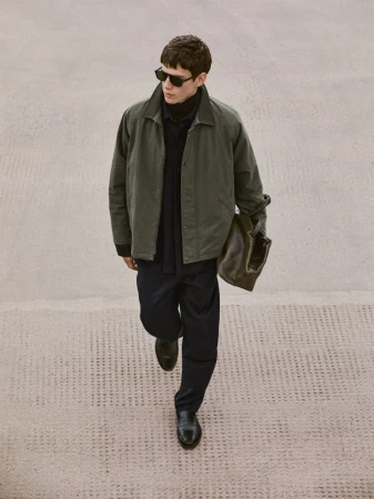Man in olive jacket, navy pants, and sunglasses walks on concrete holding a tote bag, showcasing a blend of casual and sophisticated style.
