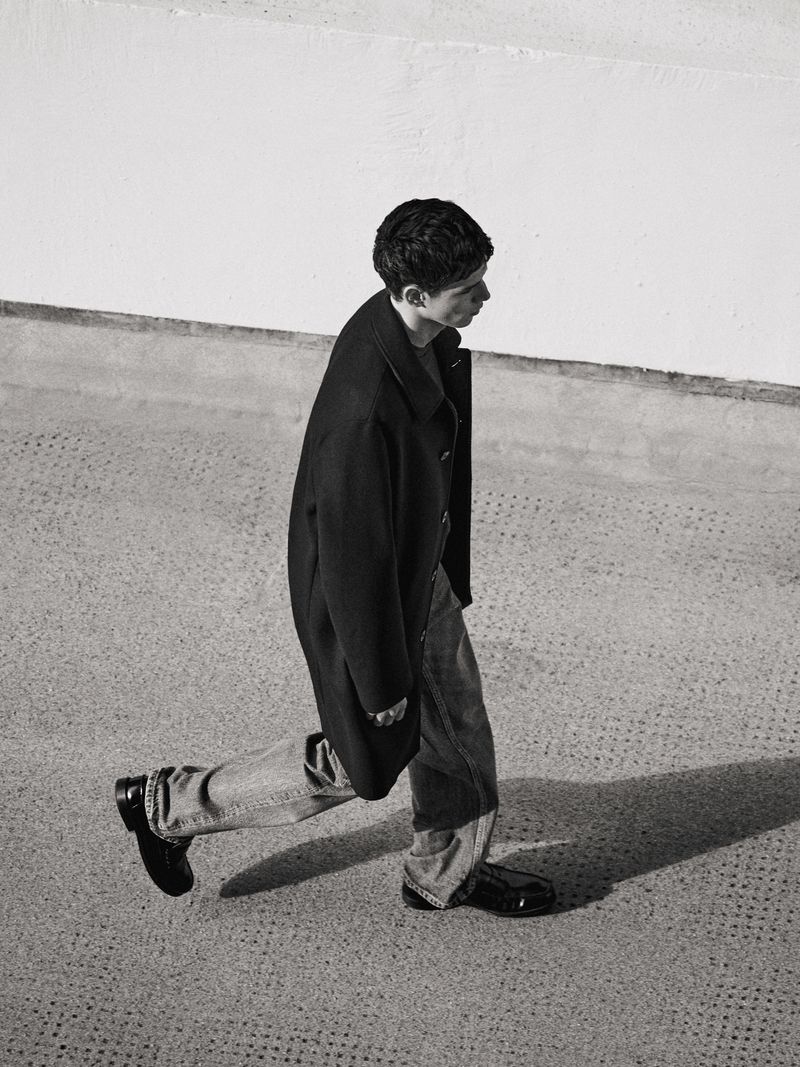 A young male model walking outdoors wearing a black overcoat, denim jeans, and dark shoes against a textured urban background.