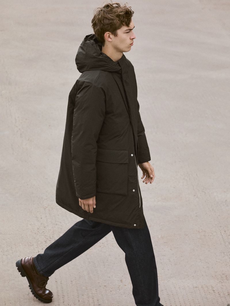 A young adult man walks outdoors on a concrete surface, wearing a dark hooded parka, dark jeans, and brown leather boots.