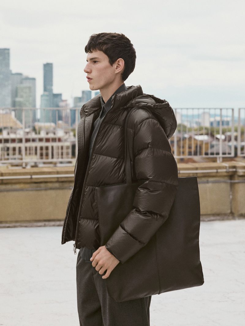A side-view medium shot of a young adult male model in a black puffer jacket, black pants, and carrying a black tote bag, against a city skyline.