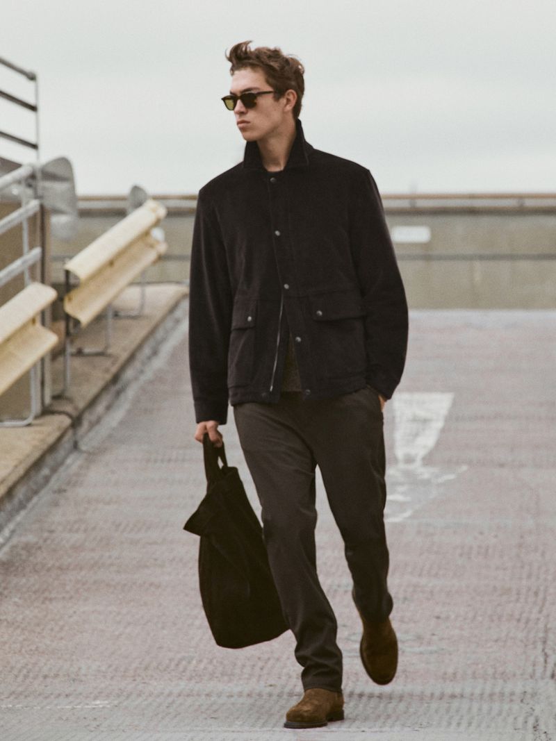 Adult male in sunglasses walks outdoors wearing jacket, pants, and shoes while carrying a black tote bag on a sunny day.