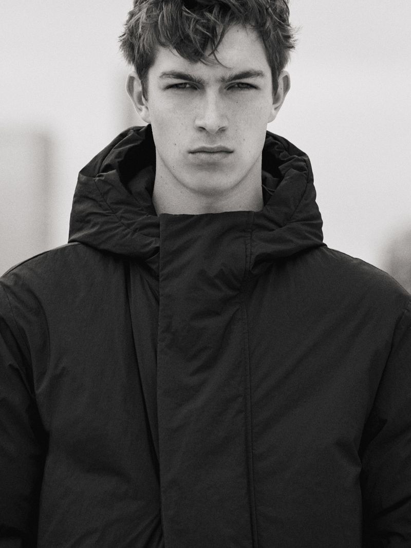 Close-up portrait of a male model wearing a dark hooded puffer jacket against a neutral backdrop.