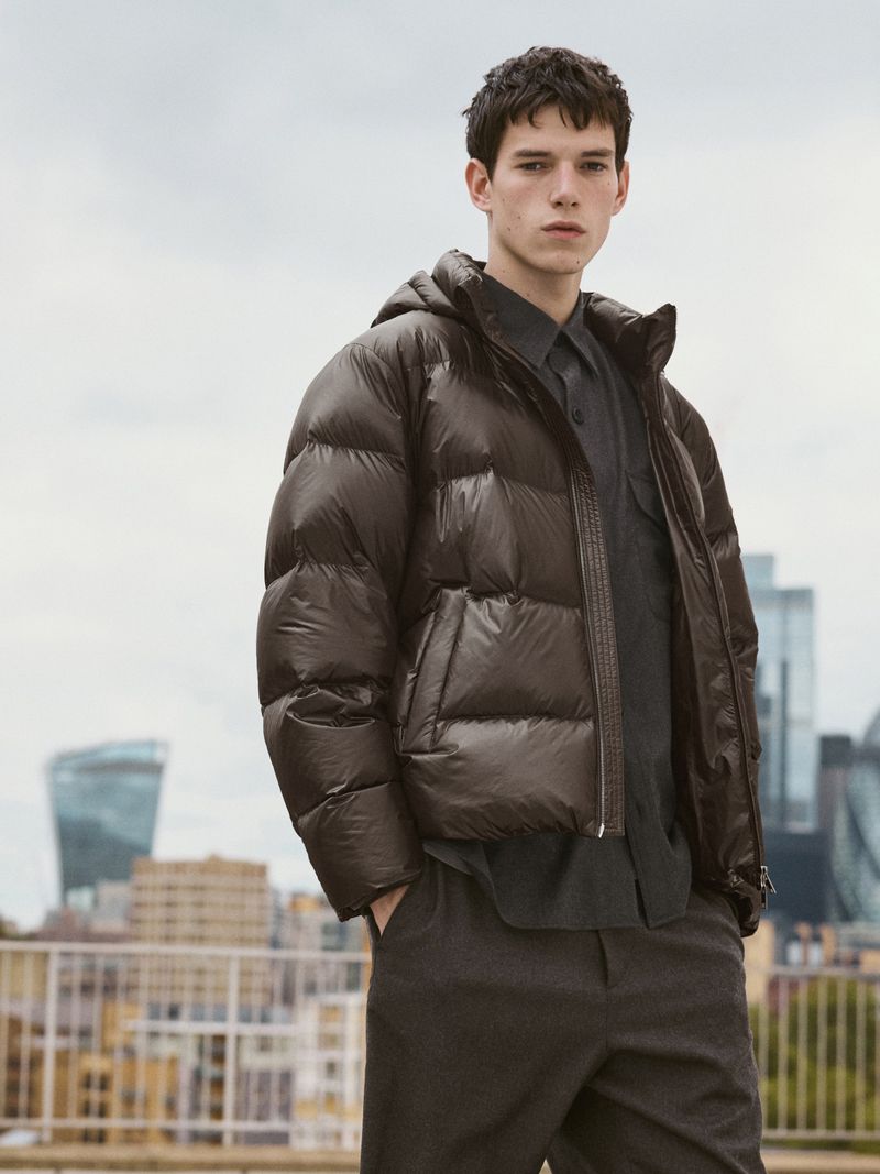 A young man in an outdoor setting wears a puffy down jacket with a button-down shirt and trousers in a fashion editorial shot.