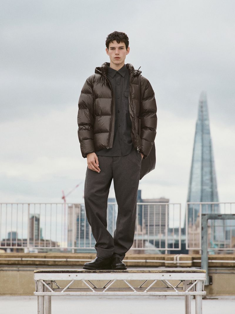 A young adult male stands on a metal platform in a grey puffer jacket and dark trousers, with an urban skyline in the background.