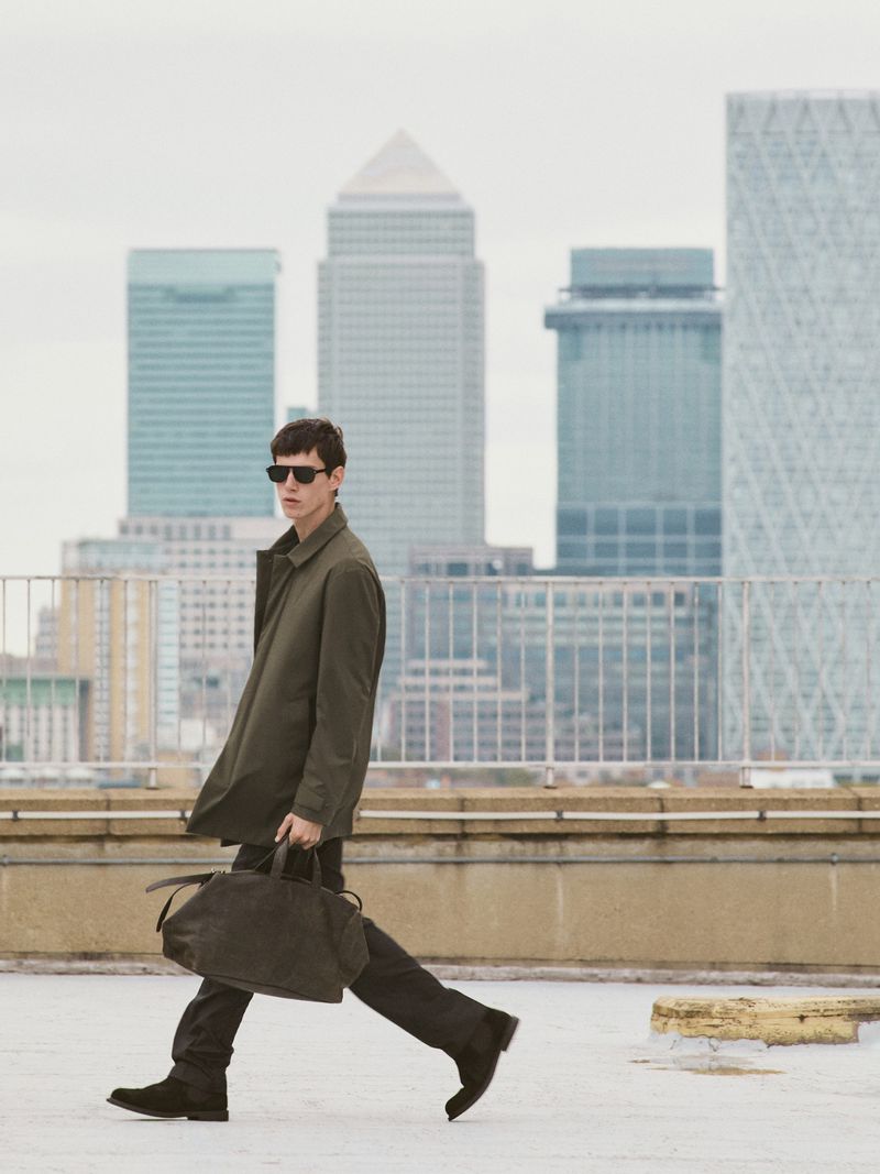 Man in coat walks with bag against city backdrop, wearing sunglasses, on concrete roof under overcast sky, showcasing fashion and architecture.