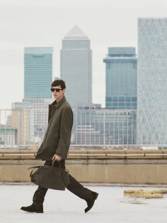 Man in coat walks with bag against city backdrop, wearing sunglasses, on concrete roof under overcast sky, showcasing fashion and architecture.